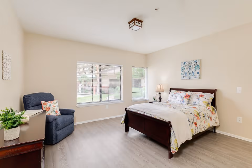 A bright and clean bedroom with a large window letting in natural light. The room features a wooden bed with floral bedding, a bedside table with a lamp, a blue armchair with a floral pillow, a wooden dresser with a potted plant, and light-colored walls and flooring.