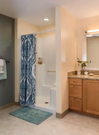 A bathroom with a walk-in shower featuring a blue and white patterned shower curtain, a green bath mat on the floor, a towel hanging on a rack, and a wooden vanity with a granite countertop, a mirror, and a small vase with flowers.