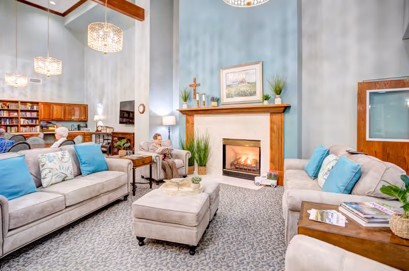 A cozy senior living facility common area with two beige sofas adorned with blue and patterned pillows, a matching ottoman, and a wooden side table with magazines. A fireplace with a wooden mantle is lit, decorated with plants, candles, a cross, and a framed picture above it. Two elderly women are seated on chairs near the fireplace. The room has light blue walls, a patterned carpet, wooden beams on the ceiling, and pendant lights. Bookshelves and a TV are visible in the background.