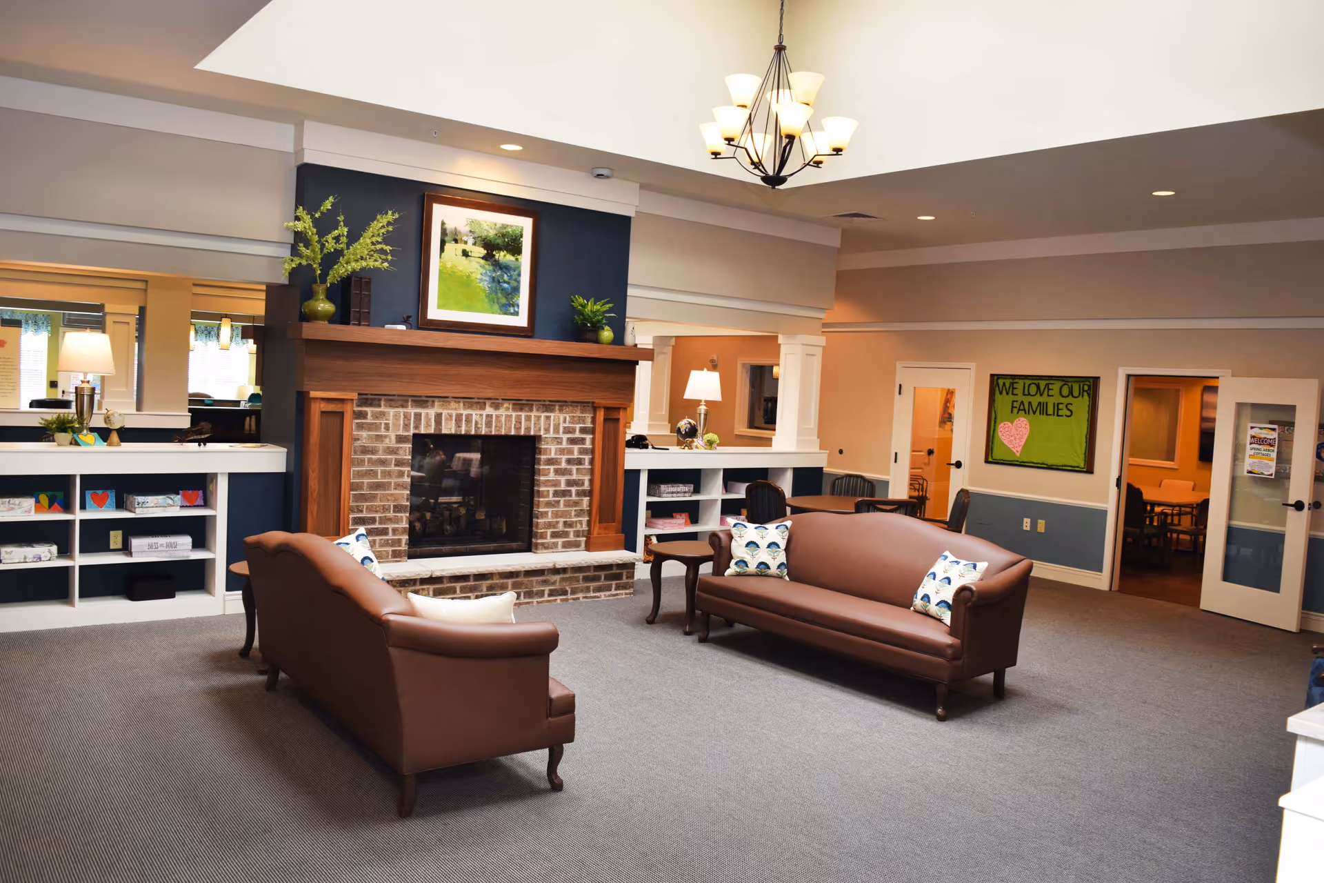 A cozy living room area in a senior living facility featuring two brown leather sofas with decorative pillows facing a brick fireplace with a wooden mantle. Above the fireplace hangs a framed painting of a green landscape. The room has carpeted floors, a chandelier with multiple lights, and built-in shelves with decorative items. A green bulletin board on the wall reads 'WE LOVE OUR FAMILIES'. There are doors leading to other rooms in the background.