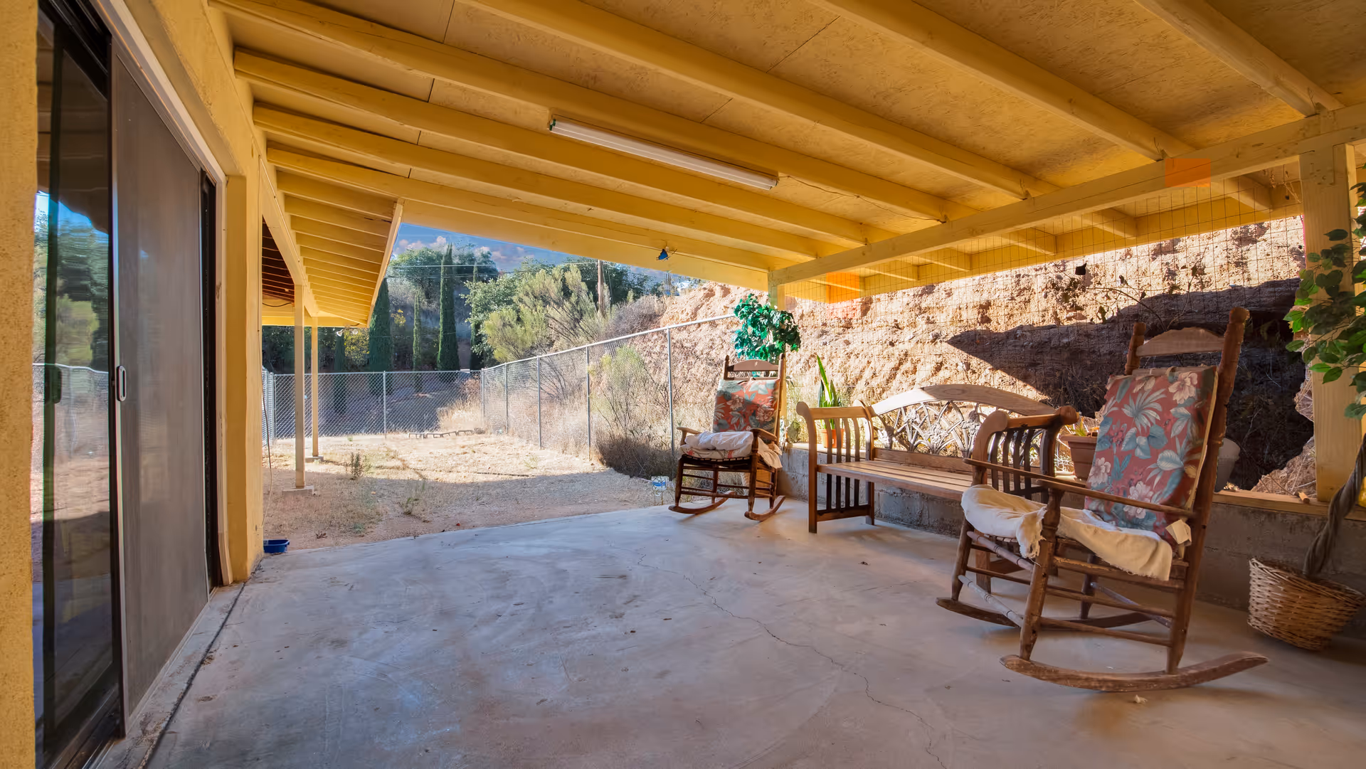 Covered outdoor patio area with a concrete floor, two wooden rocking chairs with floral cushions, a wooden bench, and a potted plant. The patio overlooks a fenced yard with dry grass and trees in the background under a clear blue sky.