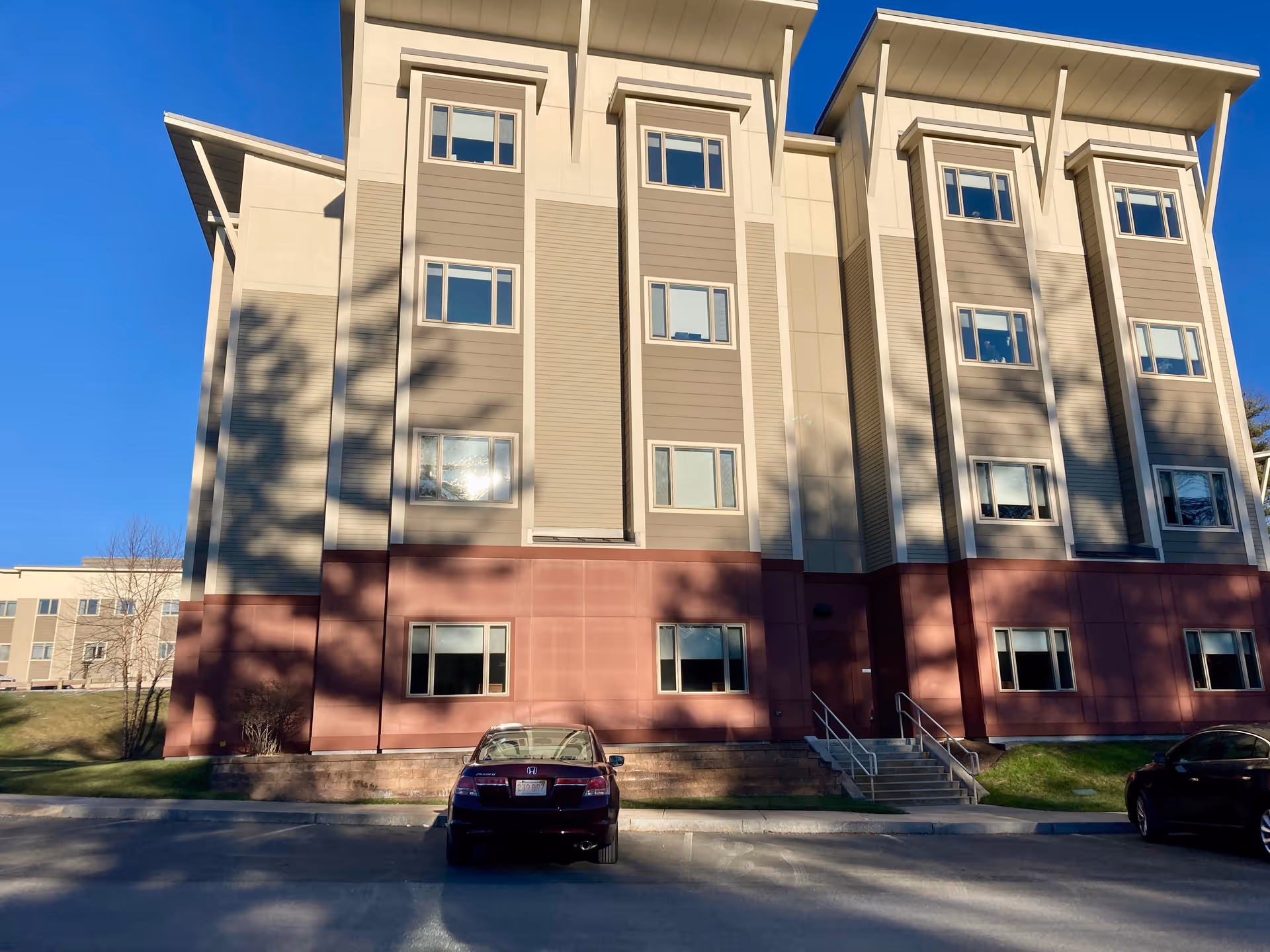 Exterior view of a multi-story building with beige and brown paneling, several windows, and a small staircase leading to a door. Two cars are parked in front of the building under a clear blue sky.