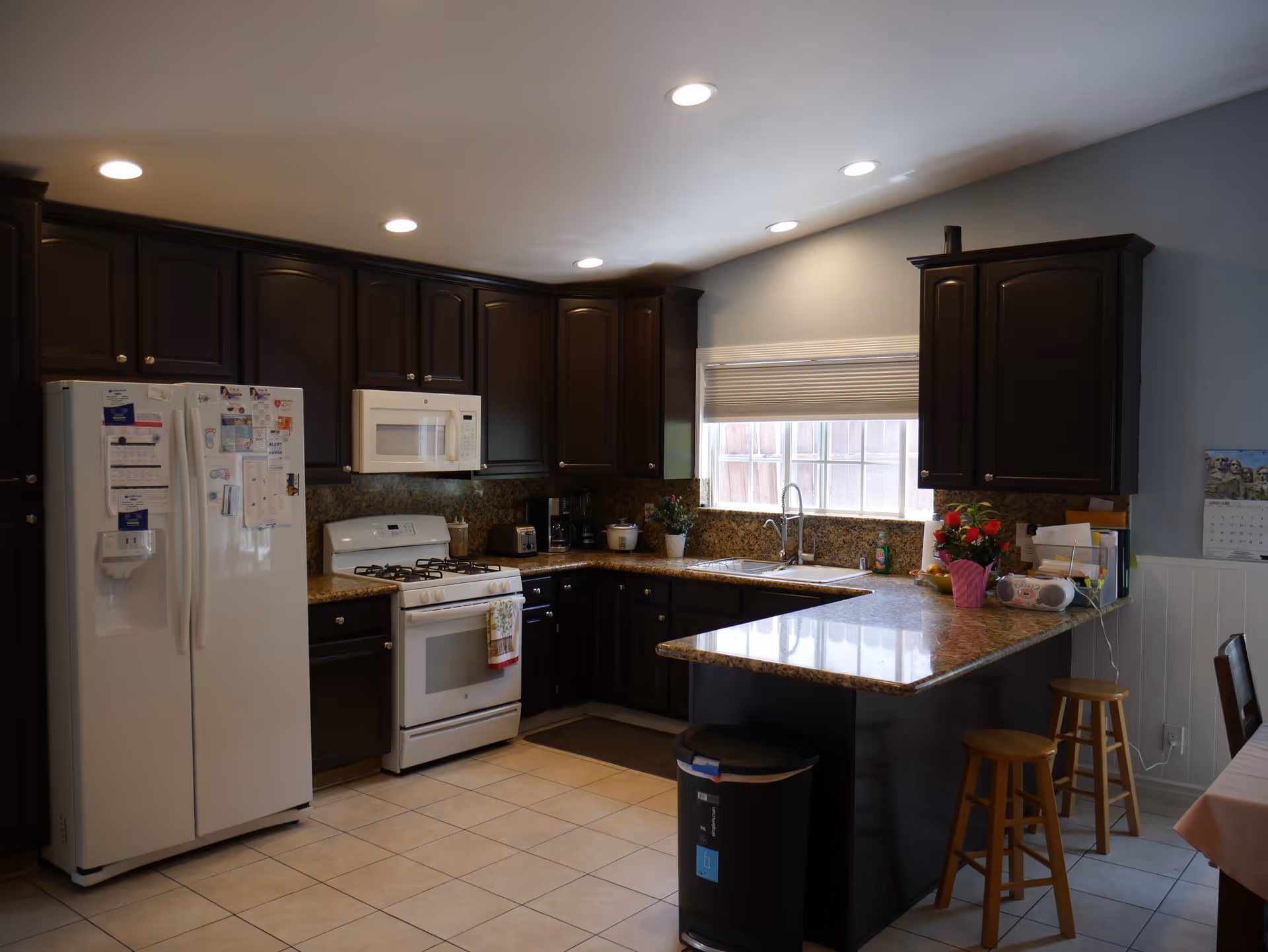 A modern kitchen with dark wood cabinets, a white refrigerator covered with magnets and papers, a white stove with a microwave above it, granite countertops, a double sink under a window with blinds, and two wooden stools at a kitchen island. The floor is tiled, and there are various kitchen appliances and decorations on the counters.