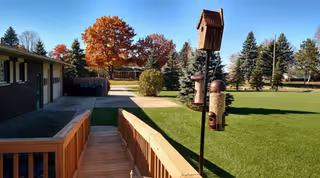 Outdoor view of a senior living facility with a wooden ramp leading to a building on the left. The scene includes a well-maintained lawn, bird feeders on poles, and trees with autumn foliage in the background under a clear blue sky.