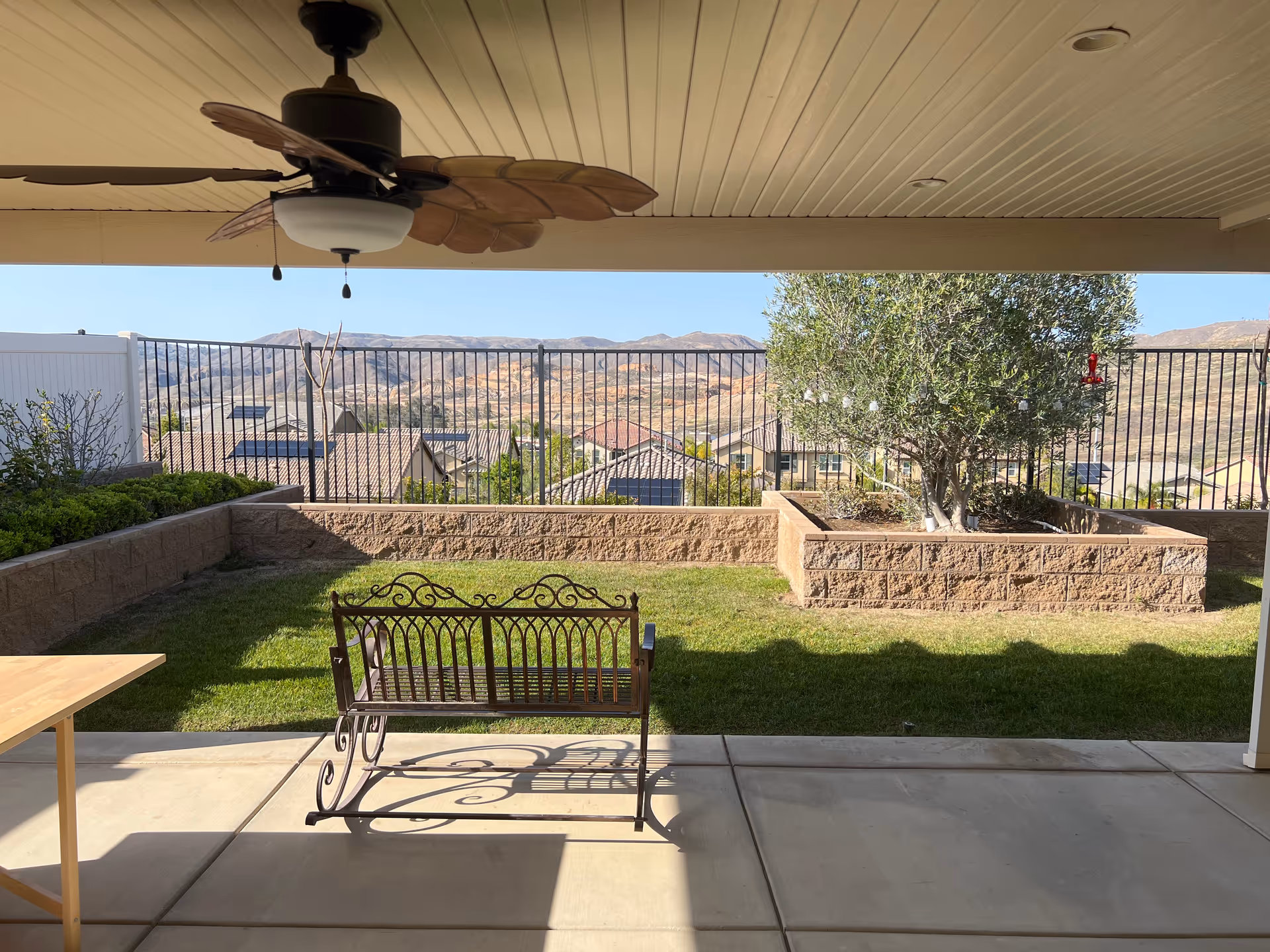 Covered patio with a ceiling fan overlooking a fenced backyard with a decorative bench, raised planters, and distant hills.