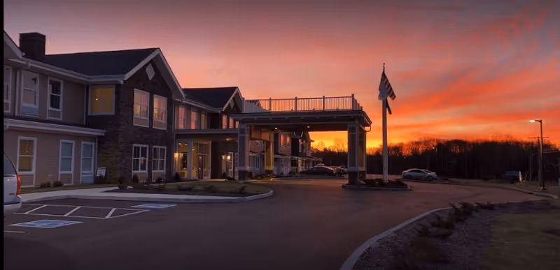 Exterior view of Legacy Living Jasper facility at sunset with a clear sky showing vibrant orange and pink hues. The building has multiple windows, a covered entrance, and a flagpole with an American flag. Several cars are parked near the entrance and along the driveway.
