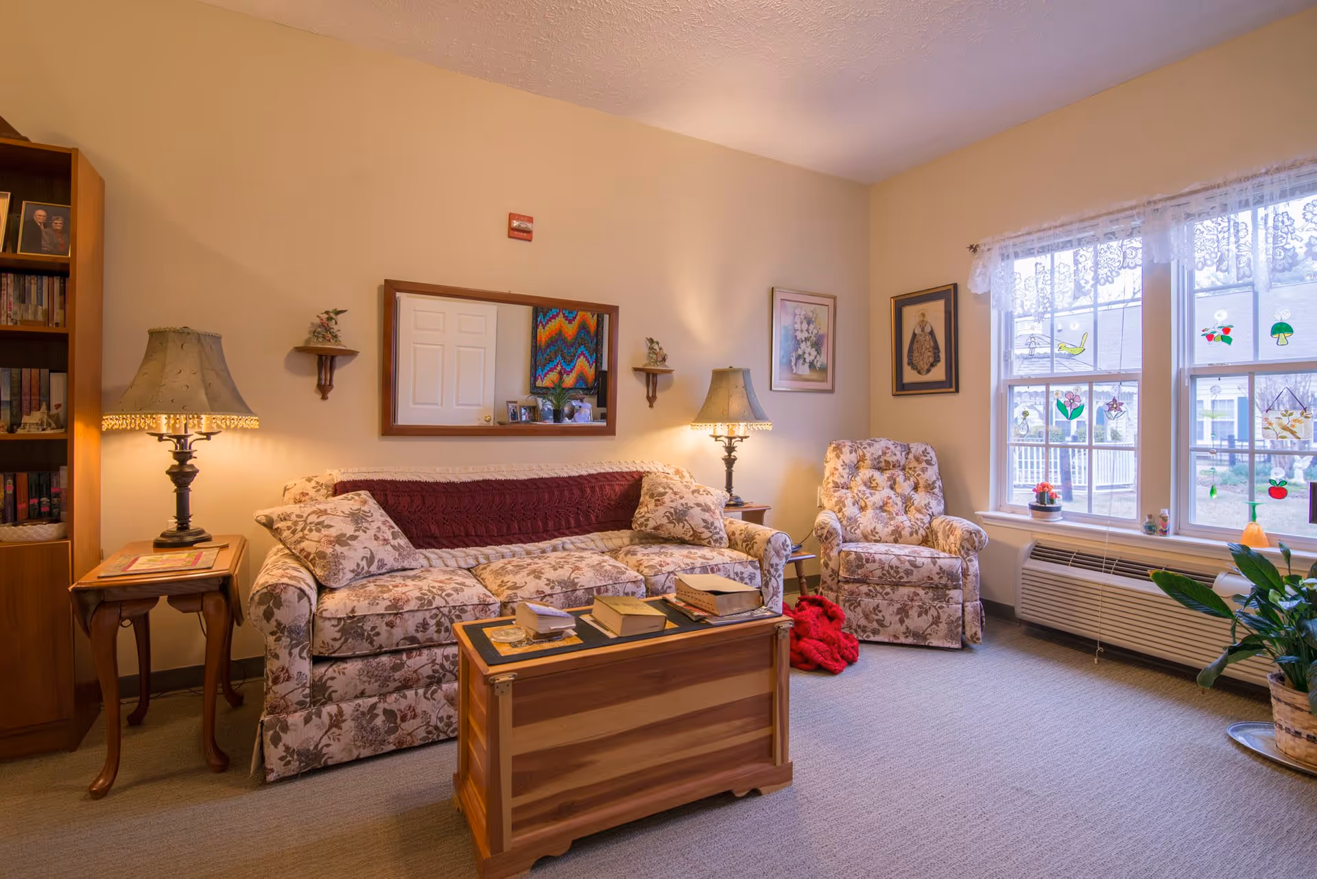 Cozy living room with a floral sofa and armchair, wooden coffee chest, lamps, bookshelf, and large windows with lace curtains.