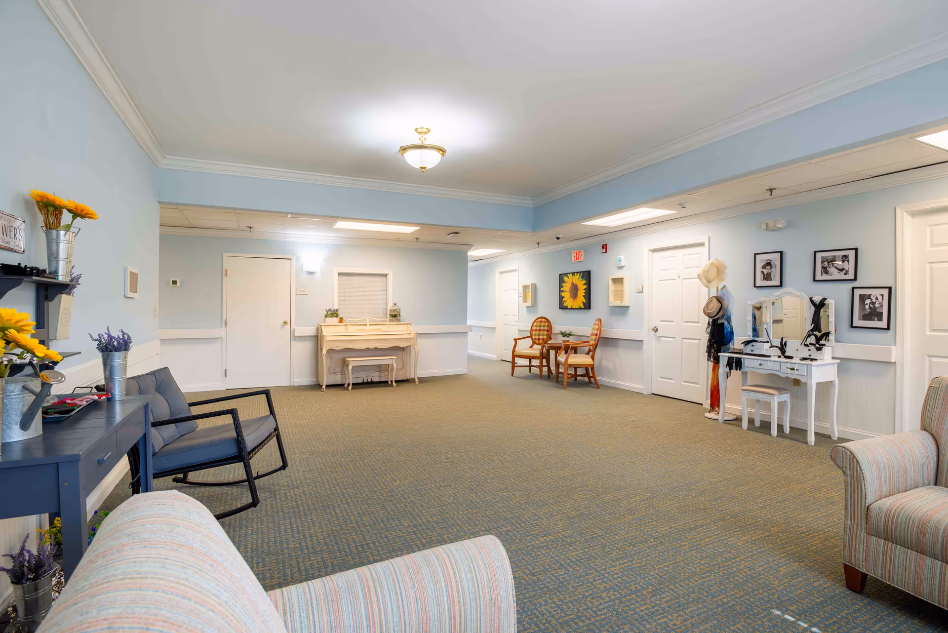 Bright, spacious senior living common room with upholstered chairs, small tables, a piano, and decorative shelving.