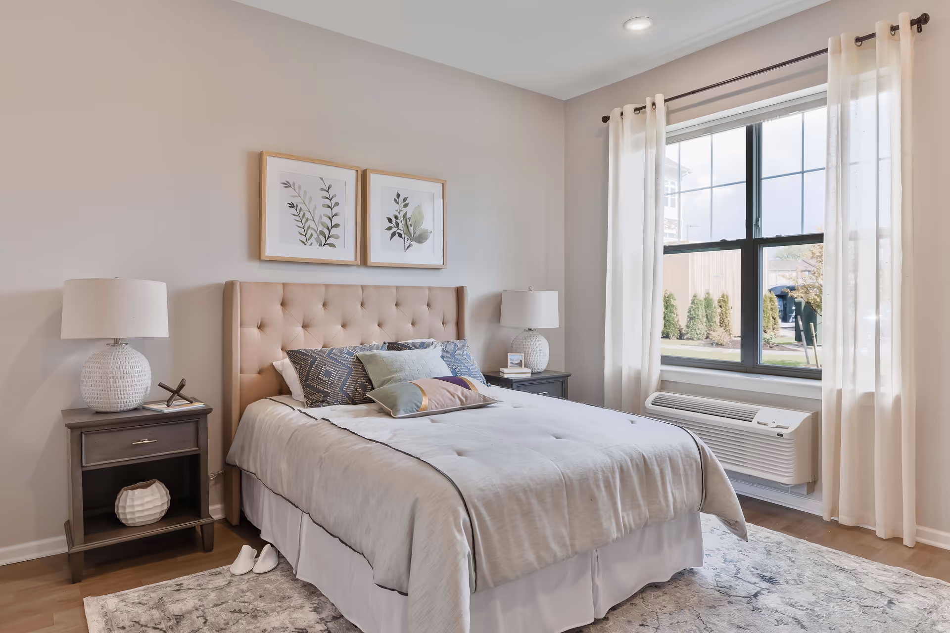 A neatly made bed with a beige tufted headboard in a bedroom. The bed has a light-colored comforter and several decorative pillows. On each side of the bed are matching dark wood nightstands with white lamps. Two framed botanical prints hang on the wall above the bed. A large window with sheer white curtains allows natural light into the room, and a heating/cooling unit is installed below the window. A patterned rug covers part of the wooden floor.