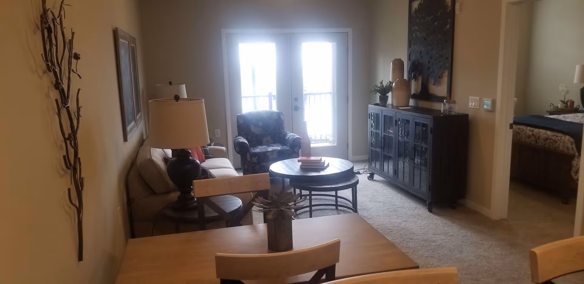 A cozy living room area with a beige sofa, a floral armchair, a round coffee table with books, and a dark wooden cabinet with decorative vases and a wall art piece above it. There is a lamp on a side table next to the sofa, and a dining table with chairs in the foreground. French doors with bright light coming through lead to an outdoor balcony. A bedroom with a bed and nightstand is visible through an open door on the right.