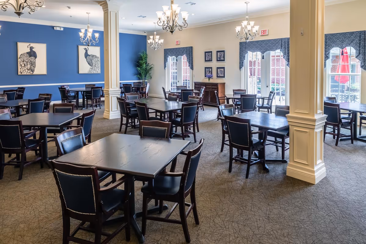 Bright dining room with multiple dark wood tables and chairs, chandeliers, a blue accent wall with artwork, and large windows.