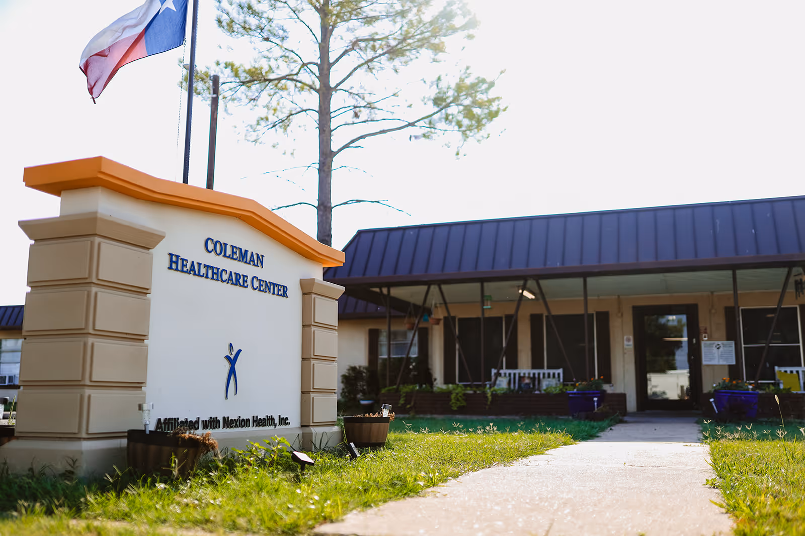 Exterior view of Coleman Healthcare Center with a sign in the foreground displaying the facility's name and affiliation with Nexion Health, Inc. A Texas state flag is flying on a pole behind the sign. The building has a dark roof and a covered entrance with a bench and potted plants.