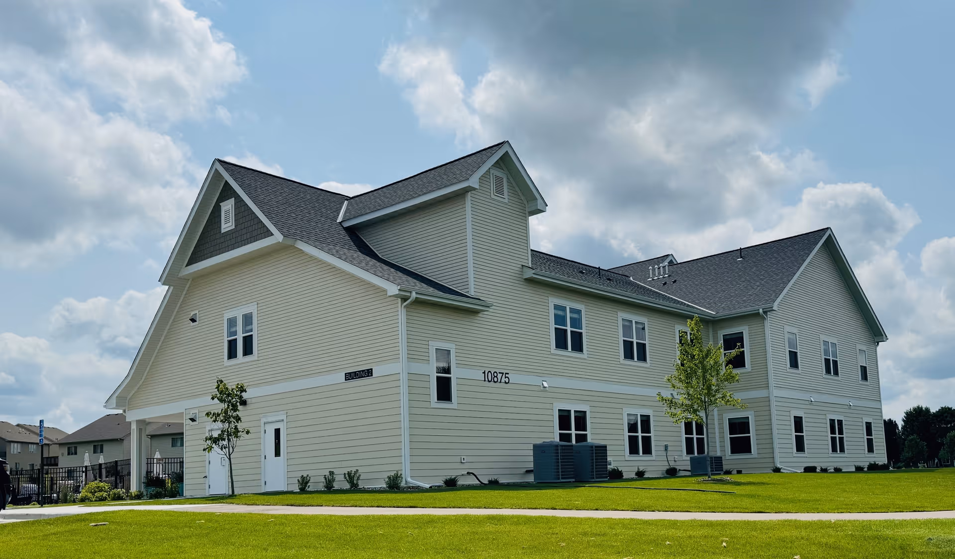 Exterior view of a two-story beige senior living facility building with multiple windows, a dark shingled roof, and a well-maintained green lawn under a partly cloudy sky.