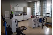 A room with medical and mobility equipment including walkers and a wheelchair, a counter with cabinets, a microwave, and a clock on the wall. The room has large windows with patterned curtains letting in natural light.