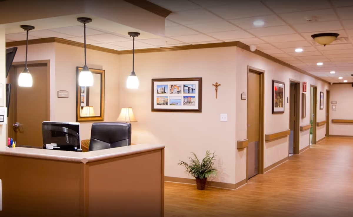 Reception desk area in a senior living facility with a computer, office chair, hanging pendant lights, framed pictures on the walls, a small potted plant, and a hallway with multiple doors and handrails.