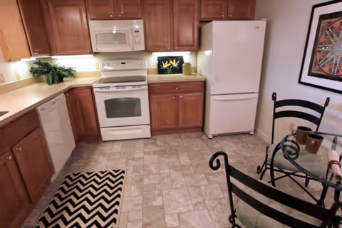 Small kitchen with white appliances, wooden cabinets, tiled floor and a small glass-top dining table.