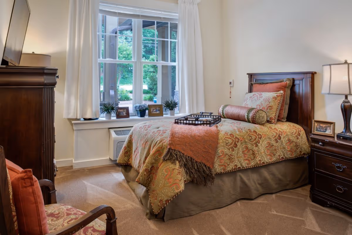 Sunlit furnished bedroom with a single bed dressed in patterned bedding, a nightstand and lamp, dresser, armchair, and a large window overlooking greenery.