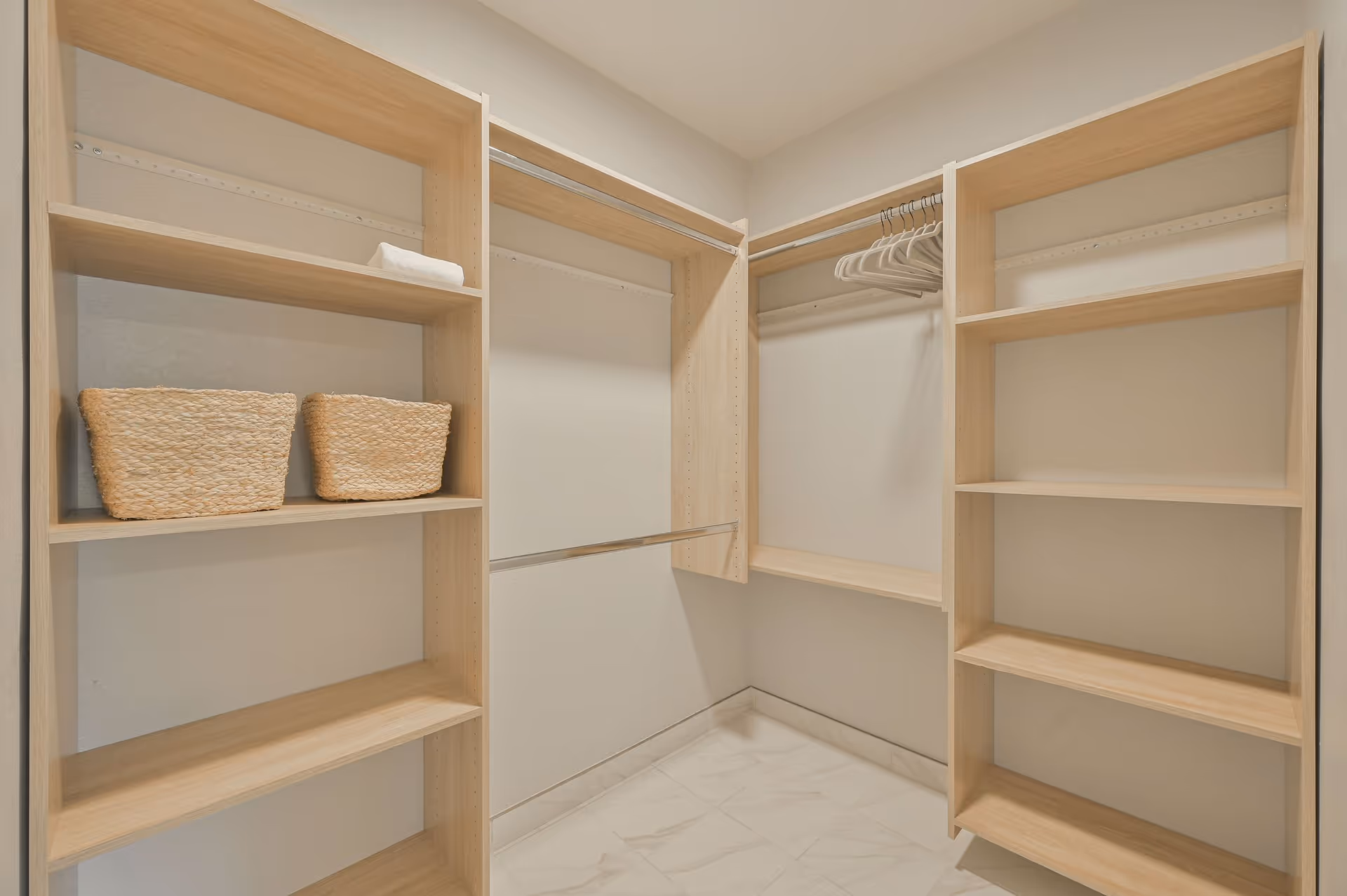 A spacious walk-in closet with light wood shelving units and hanging rods. The closet has two woven baskets on one shelf, a folded white towel on another, and several empty white hangers on a rod. The floor is tiled with a light marble pattern.