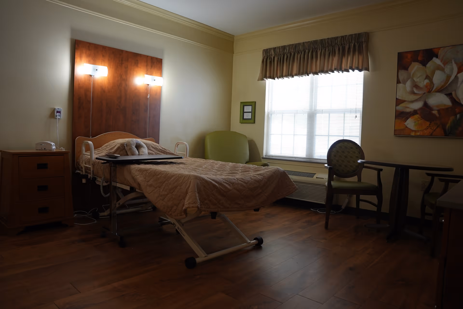 A senior living bedroom with an adjustable hospital-style bed, wall-mounted lights, a nightstand, chairs, a small table, and a window with blinds.