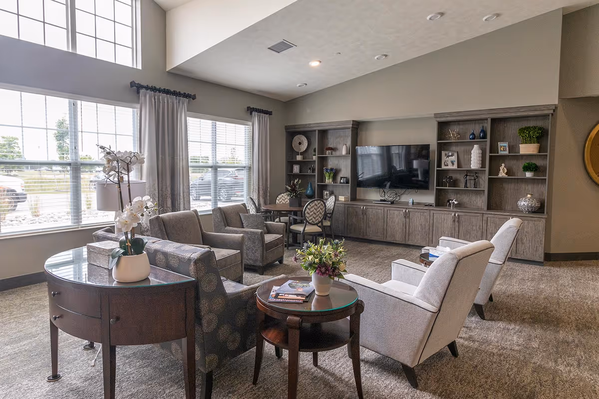 A spacious and well-lit living room area in an assisted living facility featuring multiple armchairs and a sofa arranged around a round wooden coffee table with a flower vase. Large windows with curtains allow natural light to fill the room. A large flat-screen TV is mounted on the wall above built-in wooden cabinets and shelves decorated with various ornaments and plants. The room has a neutral color palette with beige walls and carpeted flooring.