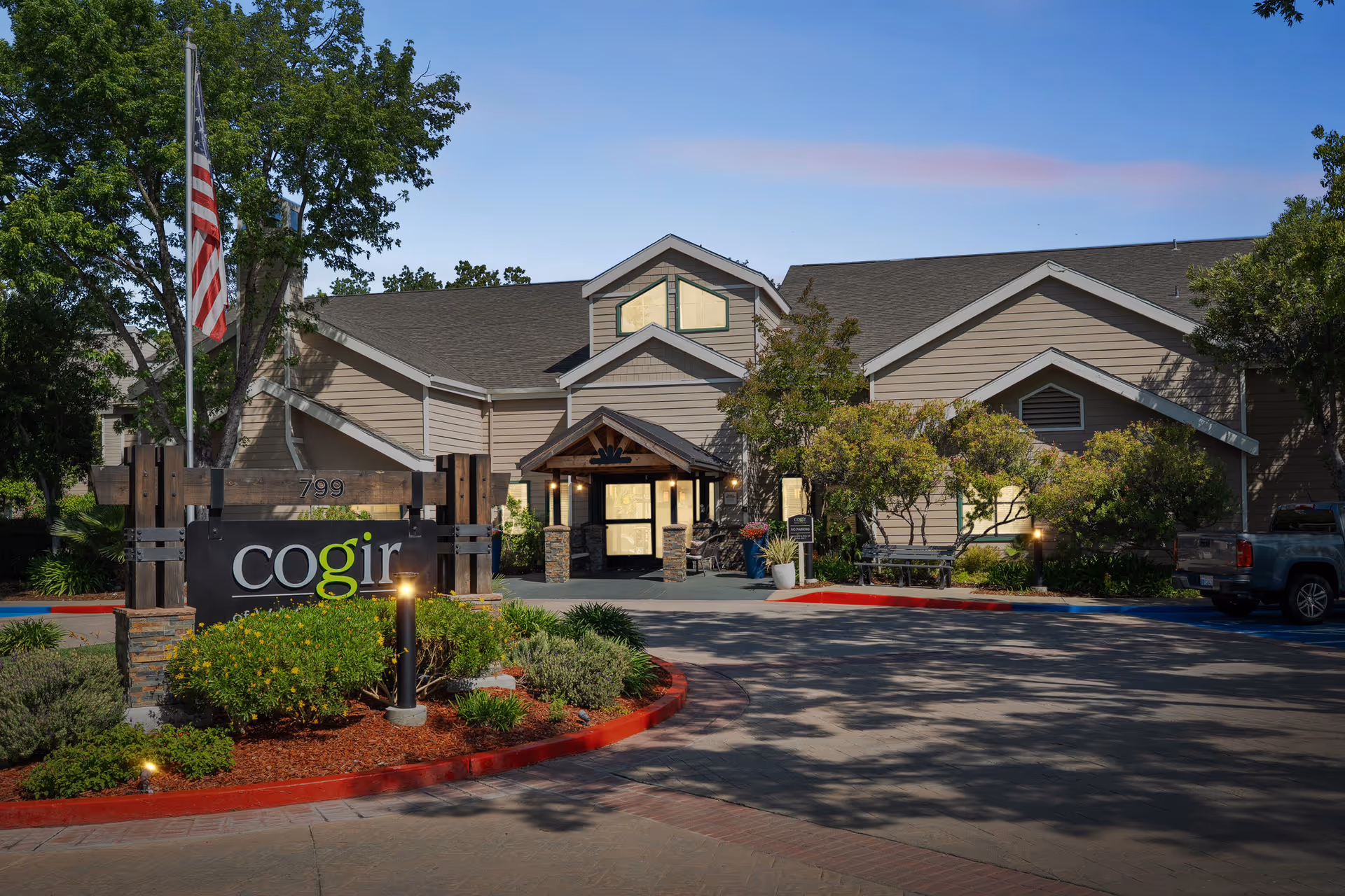 Exterior view of Cogir of Vacaville facility with a large sign displaying the name 'cogir' in front of the building. The building has a beige exterior with a peaked roof and is surrounded by trees and landscaping. An American flag is visible on a flagpole to the left of the sign. The sky is clear with a hint of pink from the setting or rising sun.