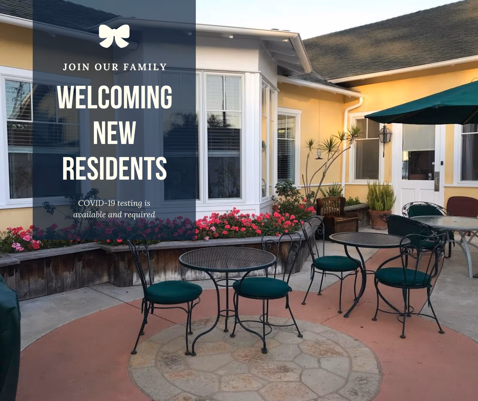 Outdoor patio area with round metal tables and chairs with green cushions, surrounded by flower beds with pink flowers and potted plants. The building exterior is painted yellow with white trim around windows and doors. A green umbrella provides shade over one of the tables. Text overlay reads 'Join our family Welcoming New Residents COVID-19 testing is available and required.'
