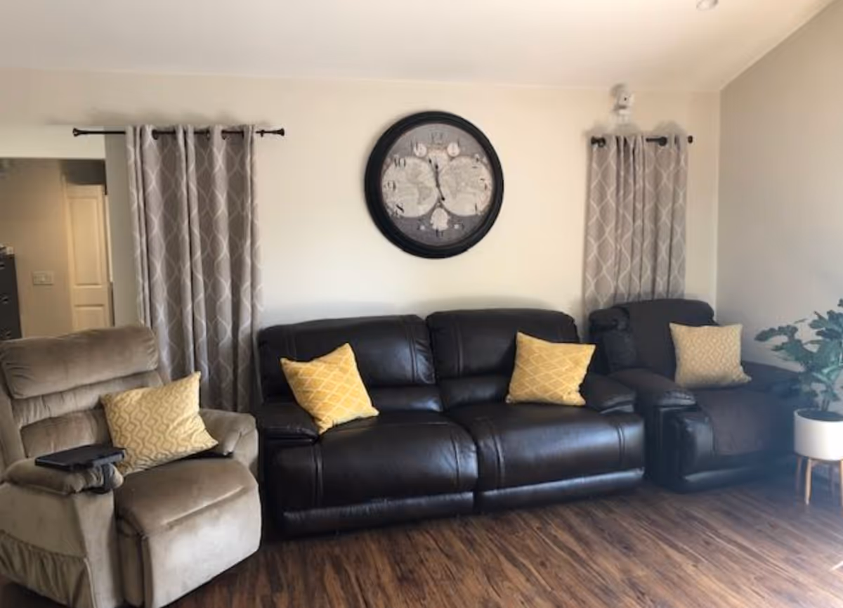 A living room with three recliner chairs, two dark brown leather and one beige fabric. Each chair has a yellow patterned pillow. Behind the chairs are two windows with gray curtains featuring a white geometric pattern. A large round wall clock with a world map design hangs on the wall between the windows. The floor is wood with a medium brown finish, and there is a potted plant in a white pot on the right side of the image.