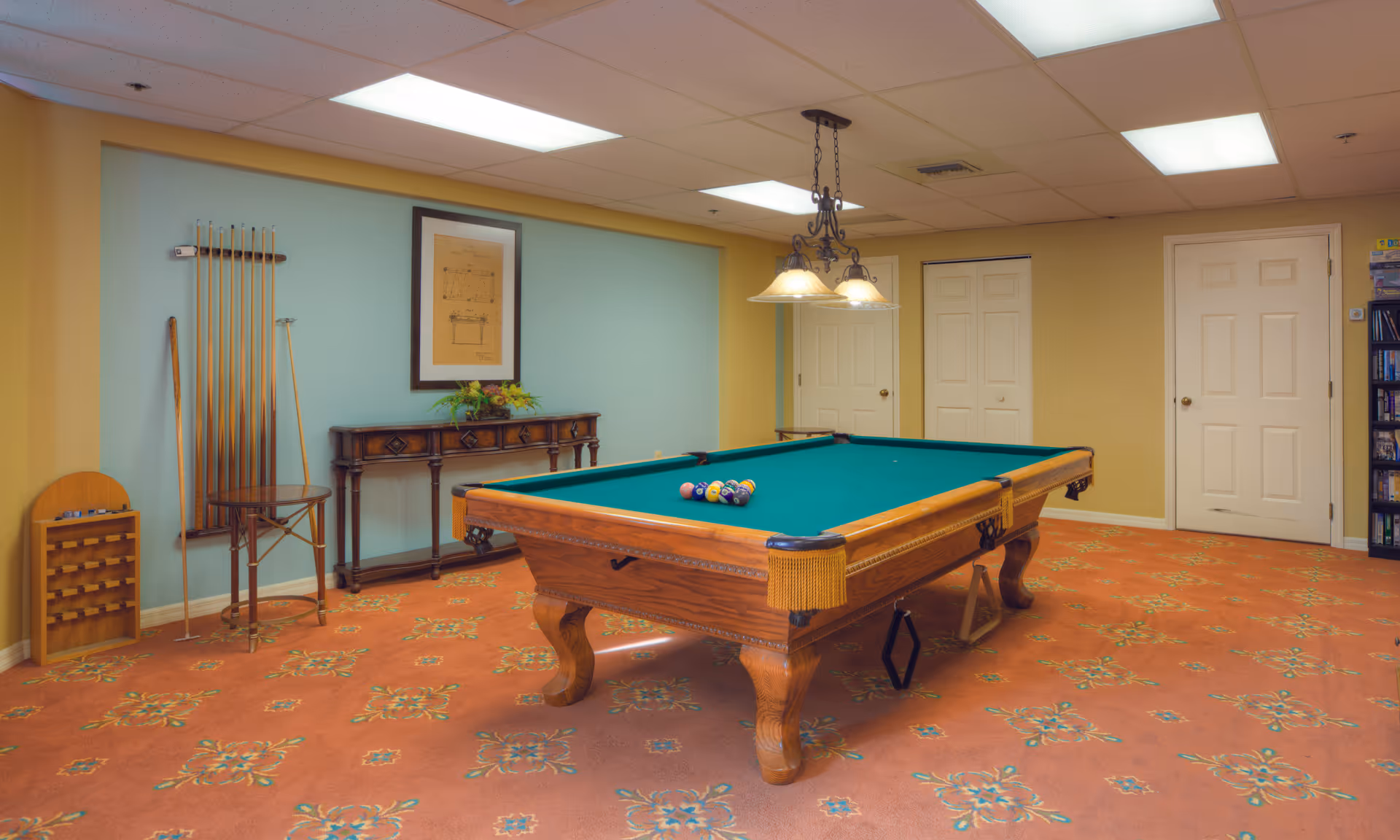 Carpeted indoor game room featuring a wooden pool table with balls racked, cue sticks on the wall, and overhead lighting.