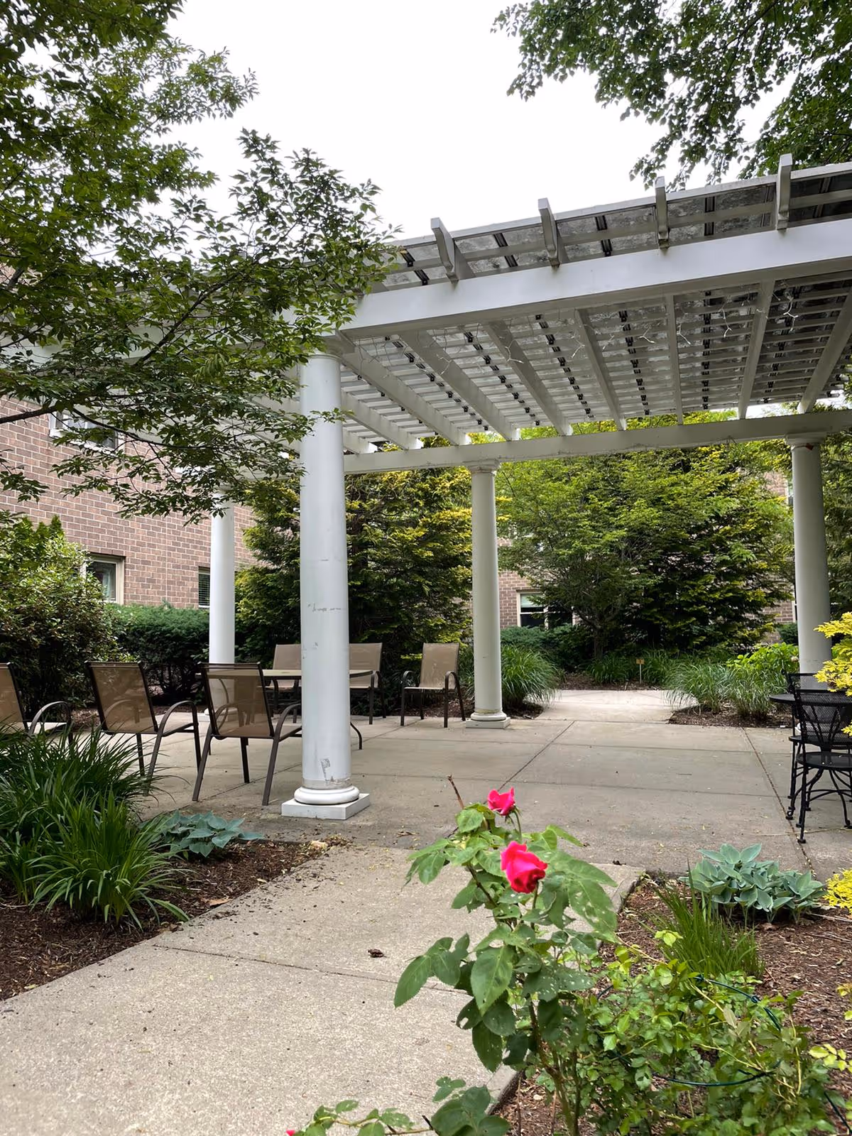 Outdoor patio area at West View Nursing & Rehabilitation Center featuring a white pergola with columns, several chairs and tables, surrounded by green bushes, trees, and flowering plants including pink roses in the foreground.