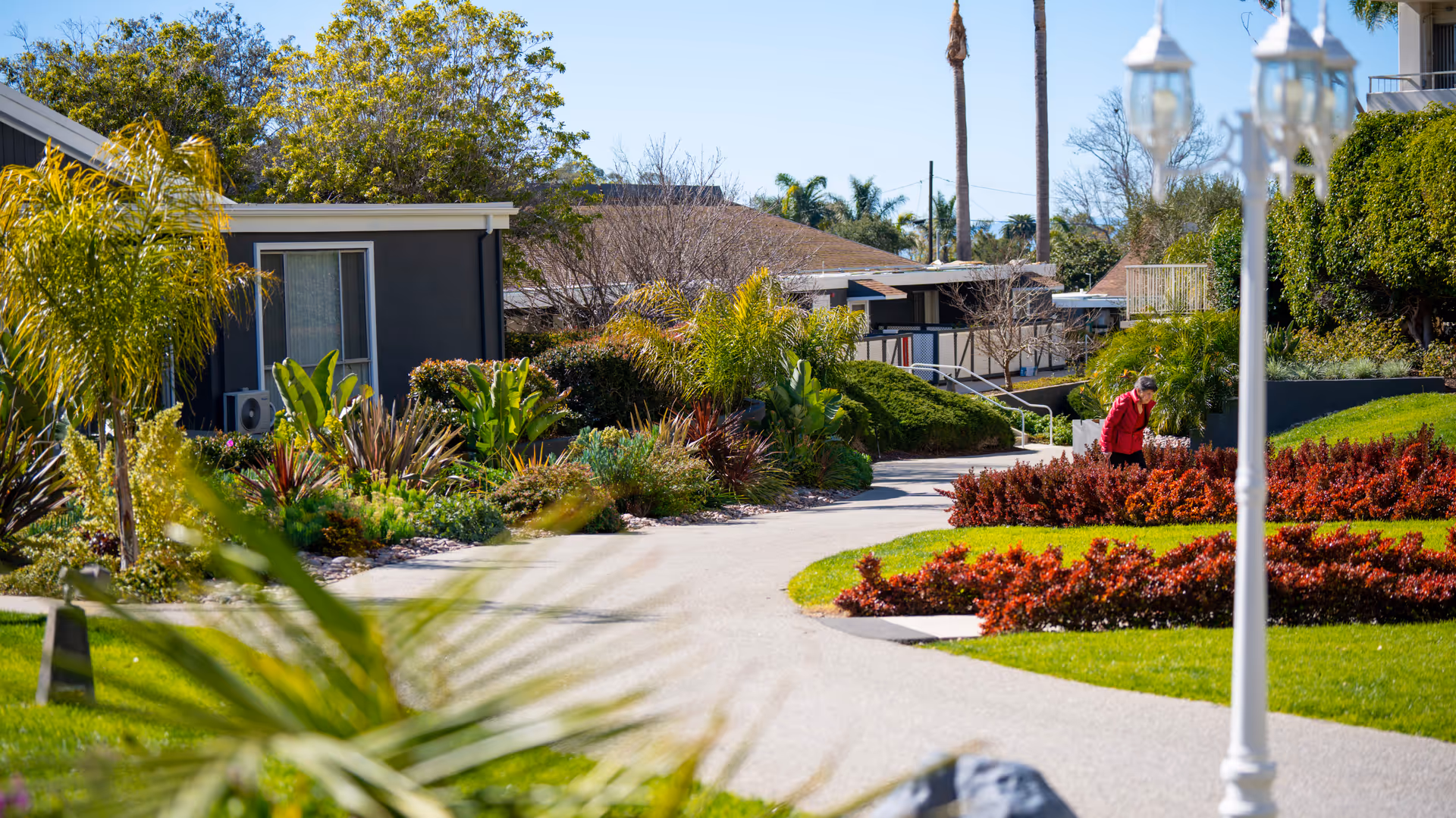 A landscaped outdoor pathway in a senior living facility with green grass, various plants, and bushes. A person wearing a red jacket is tending to the plants. Residential buildings and palm trees are visible in the background under a clear blue sky.