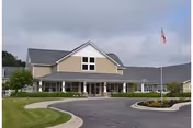 Exterior view of a single-story senior living facility building with beige siding and a gray roof. The building has a covered entrance with white columns and several windows. There is a curved driveway in front, landscaped with grass and small bushes, and an American flag on a flagpole near the entrance.