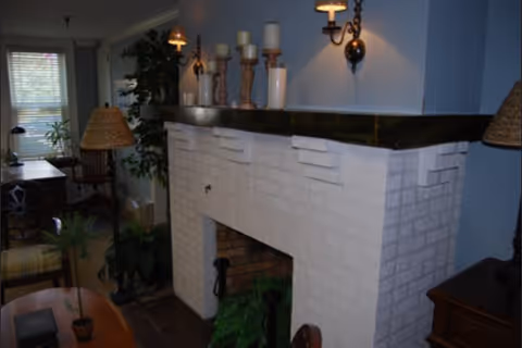 Cozy living room with a white painted brick fireplace mantel topped with candles and wall sconces, surrounded by plants and seating.