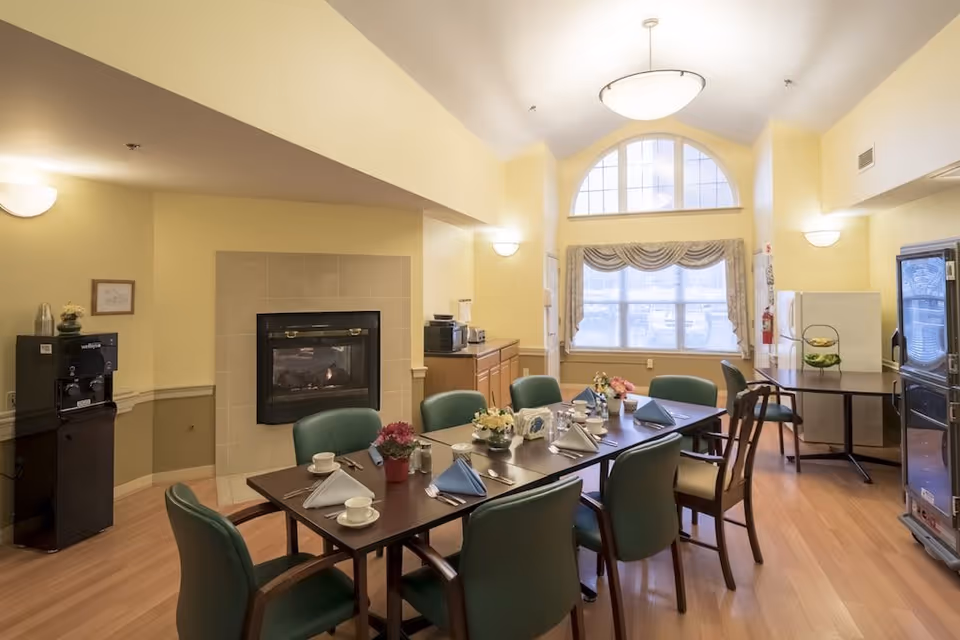 A dining room in an assisted living facility with a long table set for a meal, including cups, plates, napkins, and flowers. The room has green chairs around the table, a fireplace on one wall, a large window with curtains, and kitchen appliances like a refrigerator and microwave in the background.