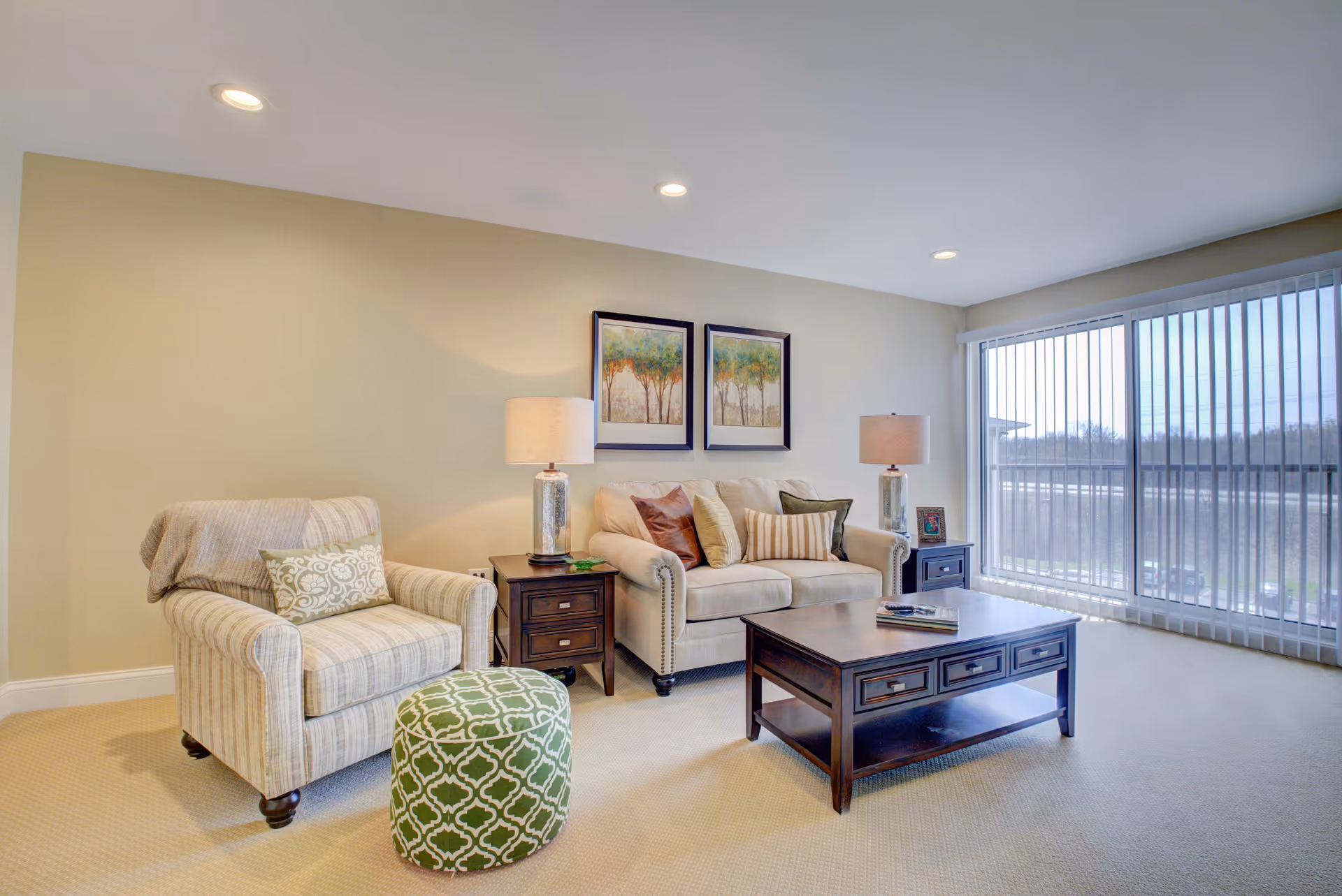 A bright living room with a beige armchair and a matching sofa adorned with multiple cushions. There are two side tables with lamps on either side of the sofa. A dark wooden coffee table is placed in front of the sofa. Large sliding glass doors with vertical blinds allow natural light to fill the room. Two framed paintings of trees hang on the wall above the sofa.