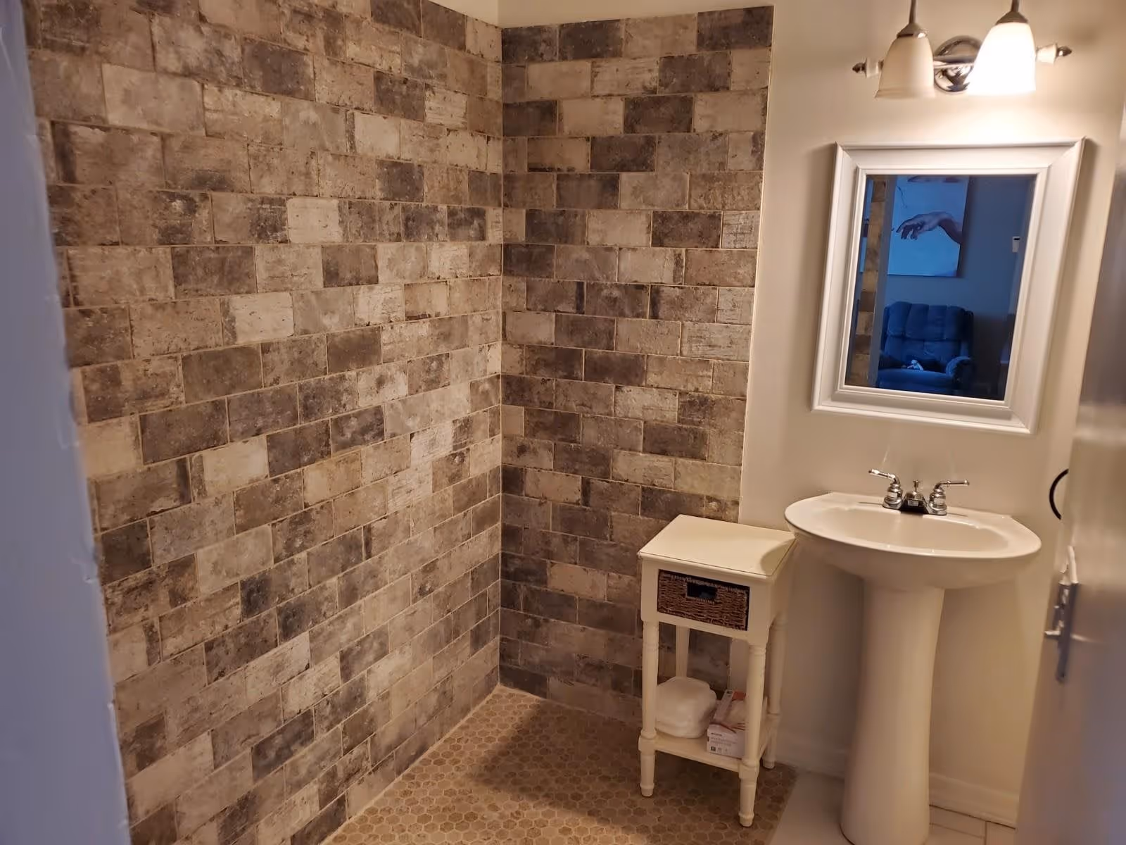 A bathroom with a tiled shower area featuring brown and beige rectangular tiles. There is a white pedestal sink with a chrome faucet, a small white side table with a wicker drawer holding some items, and a mirror above the sink reflecting part of a room with a blue armchair and a painting of a hand.