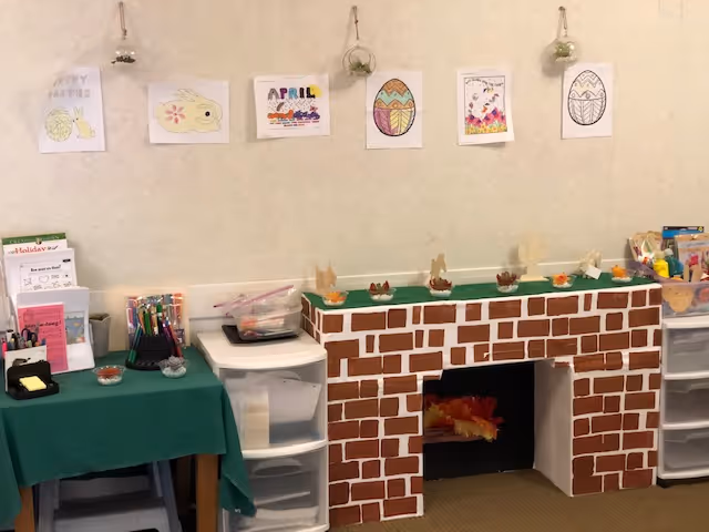 A room corner decorated with a faux brick fireplace made of painted cardboard, topped with small Easter-themed decorations. Above the fireplace, six children's drawings featuring Easter eggs, a bunny, and the word 'April' are hung on the wall. To the left, a table covered with a green cloth holds various office supplies and papers, and to the right, plastic drawers contain more supplies.