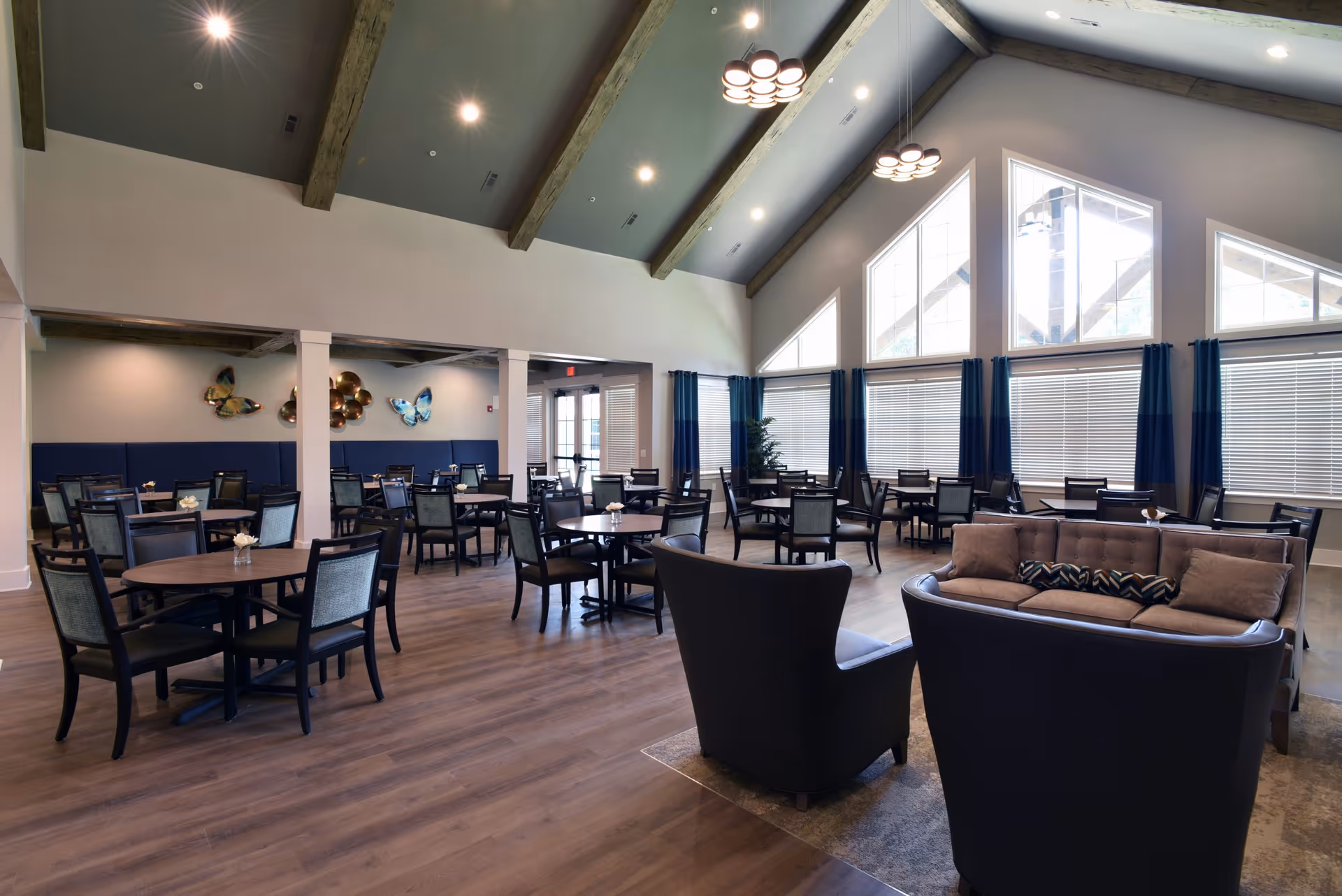 A spacious senior living dining room with multiple round tables and chairs arranged neatly. The room features high vaulted ceilings with exposed wooden beams, large windows with blinds and blue curtains, and modern light fixtures. There is also a seating area with a brown sofa and two black armchairs on a rug in the foreground. Wall decorations include butterfly art pieces.