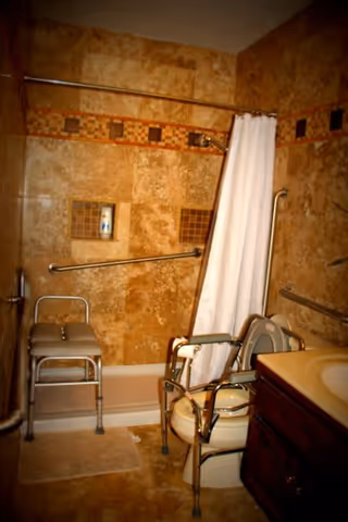 A bathroom designed for elderly or disabled use featuring a shower area with grab bars, a shower chair, a white shower curtain, and a raised toilet seat with armrests. The walls are tiled with a beige and brown mosaic pattern, and there is a sink with a wooden cabinet visible on the right side.