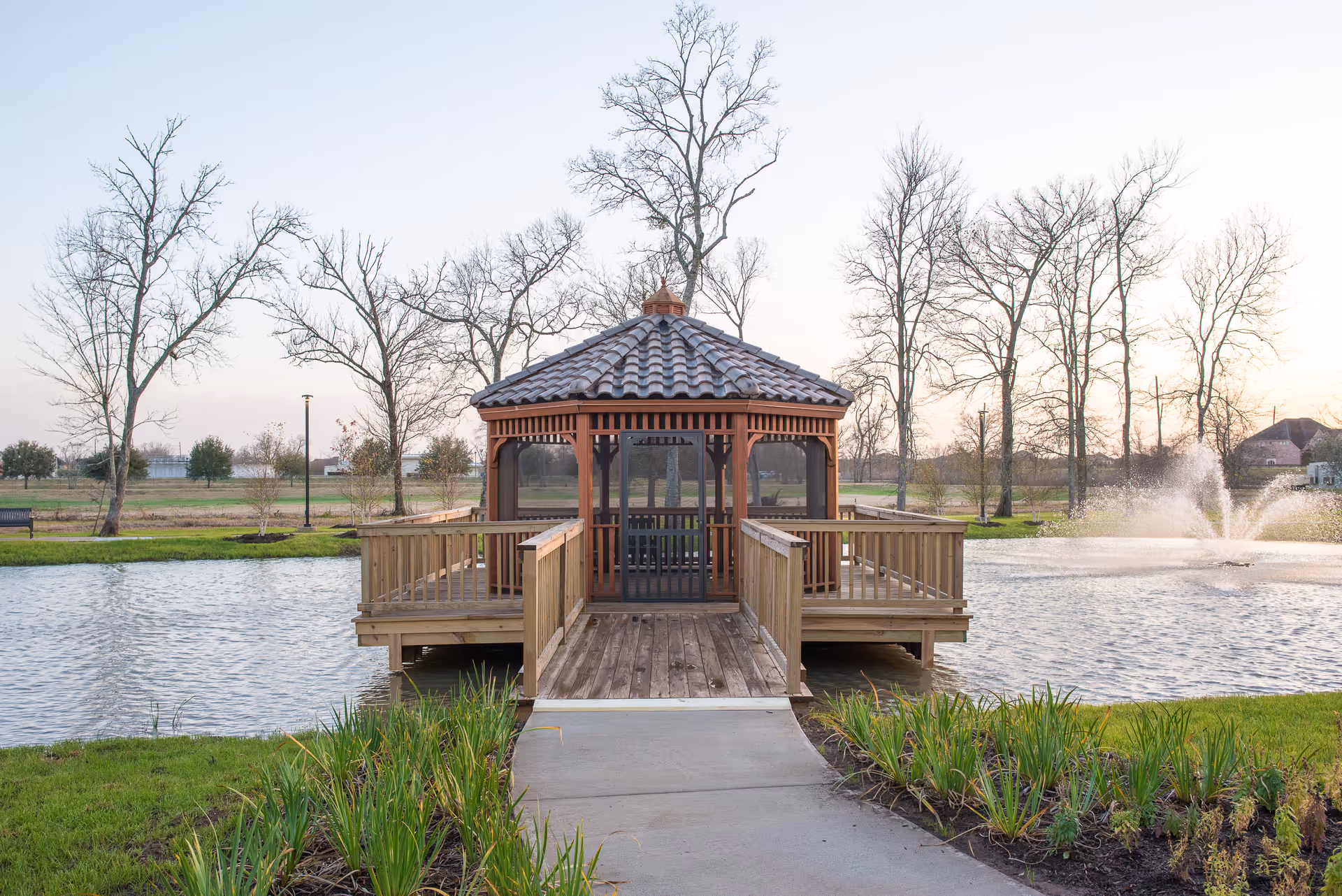 A wooden gazebo with a tiled roof situated on a small pier extending over a pond with a fountain, surrounded by grass and leafless trees under a clear sky.