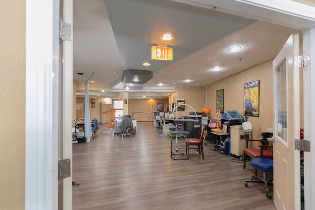 Interior view of a rehabilitation or therapy room with wooden flooring, various chairs, desks with office supplies, medical equipment, and motivational posters on the walls. The room is well-lit with ceiling lights and has an exit sign above the doorway.