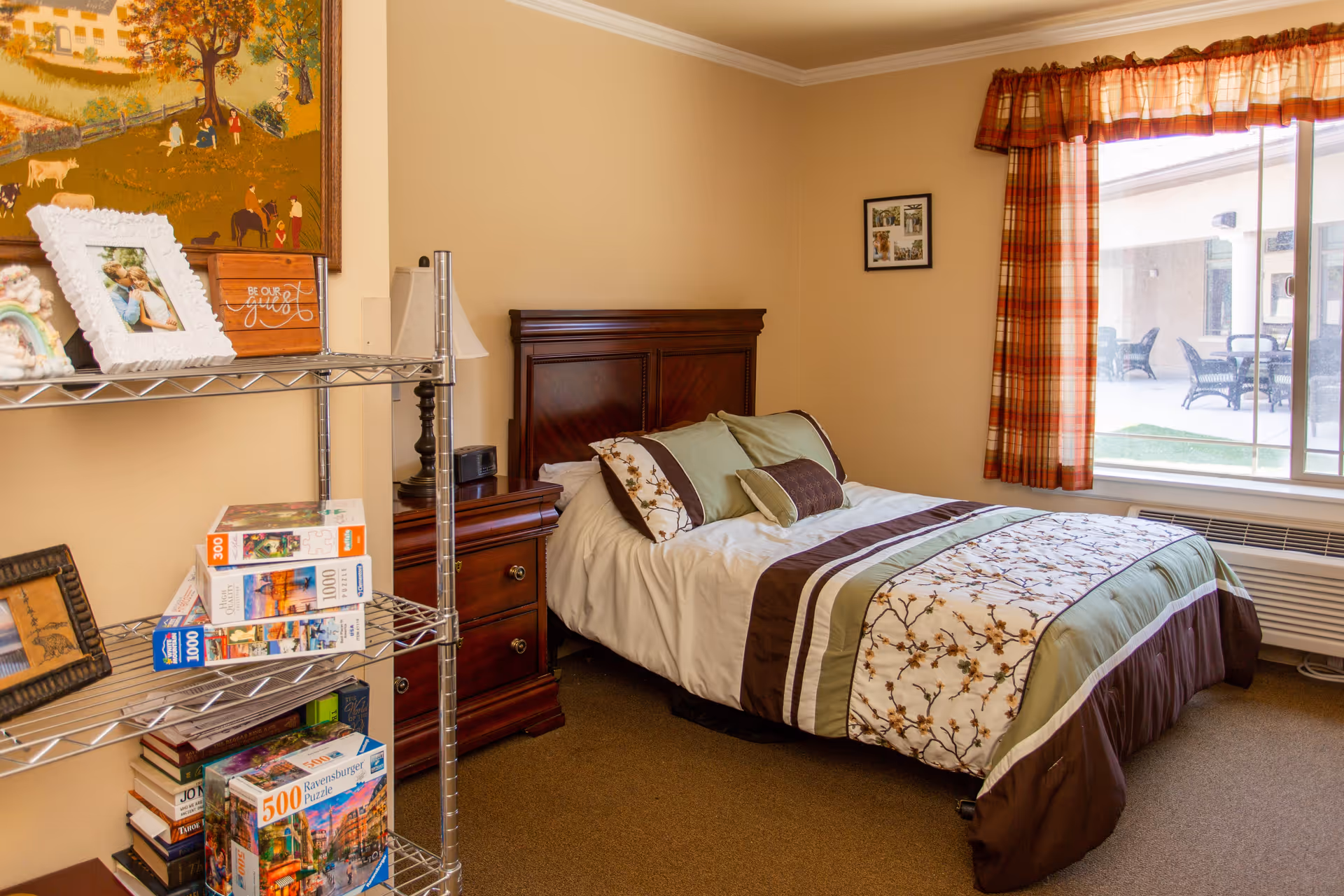 A cozy bedroom with a neatly made bed featuring a floral and striped comforter in shades of green, brown, and white. Next to the bed is a wooden nightstand with a lamp and a clock. A metal shelving unit holds several puzzle boxes and framed photos. A large window with plaid curtains lets in natural light and shows an outdoor patio area with chairs and tables.