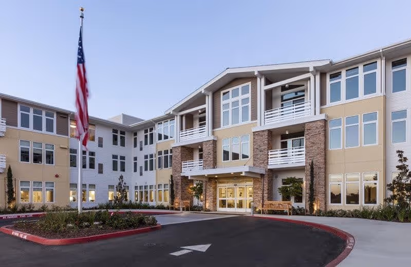 Exterior view of a modern three-story senior living facility building with large windows, balconies, and a covered entrance. There is a circular driveway with a landscaped island featuring an American flagpole in front of the building.
