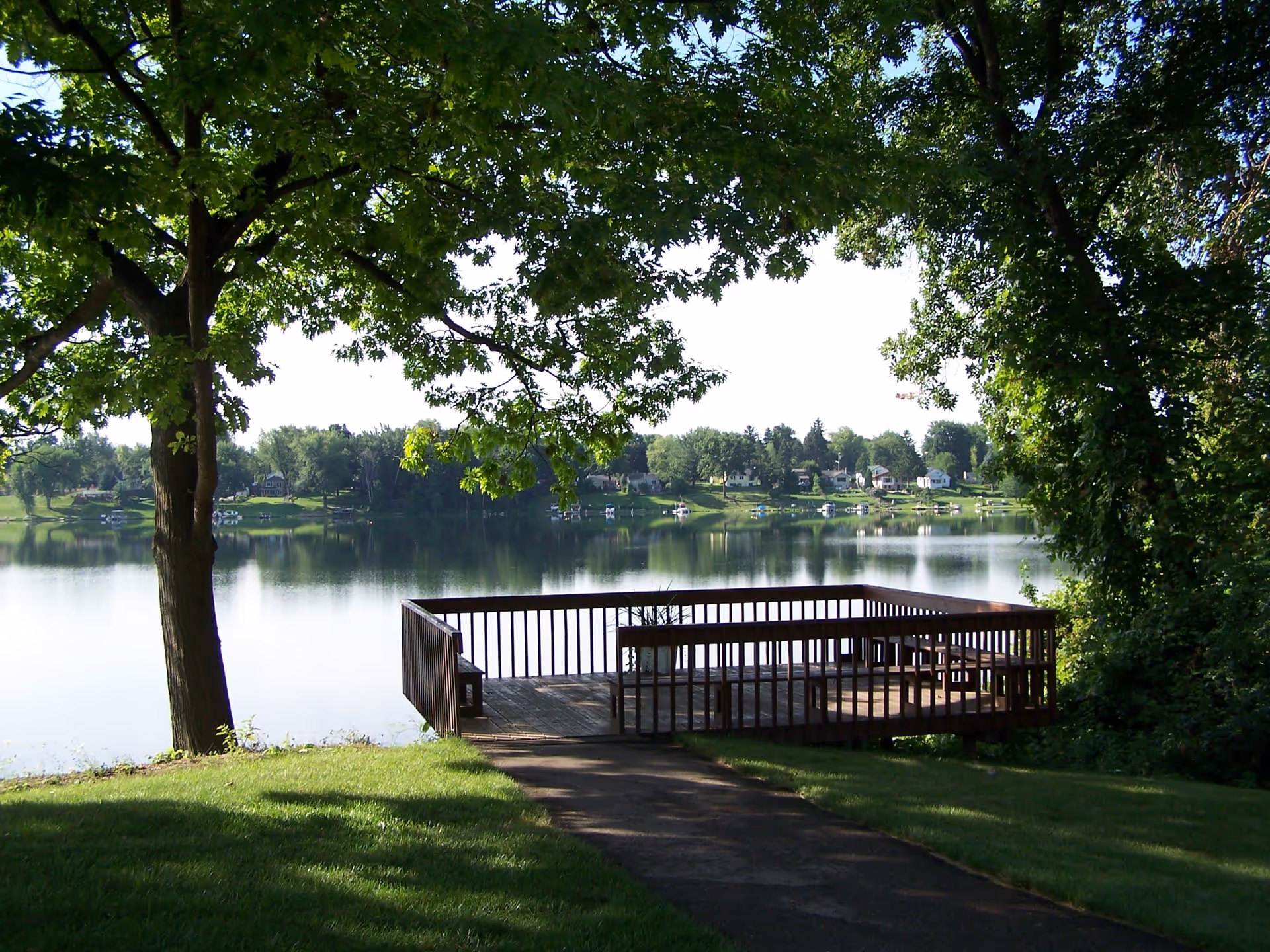A wooden deck with benches overlooking a calm lake, surrounded by green trees and grass, with houses visible across the water under a clear sky.