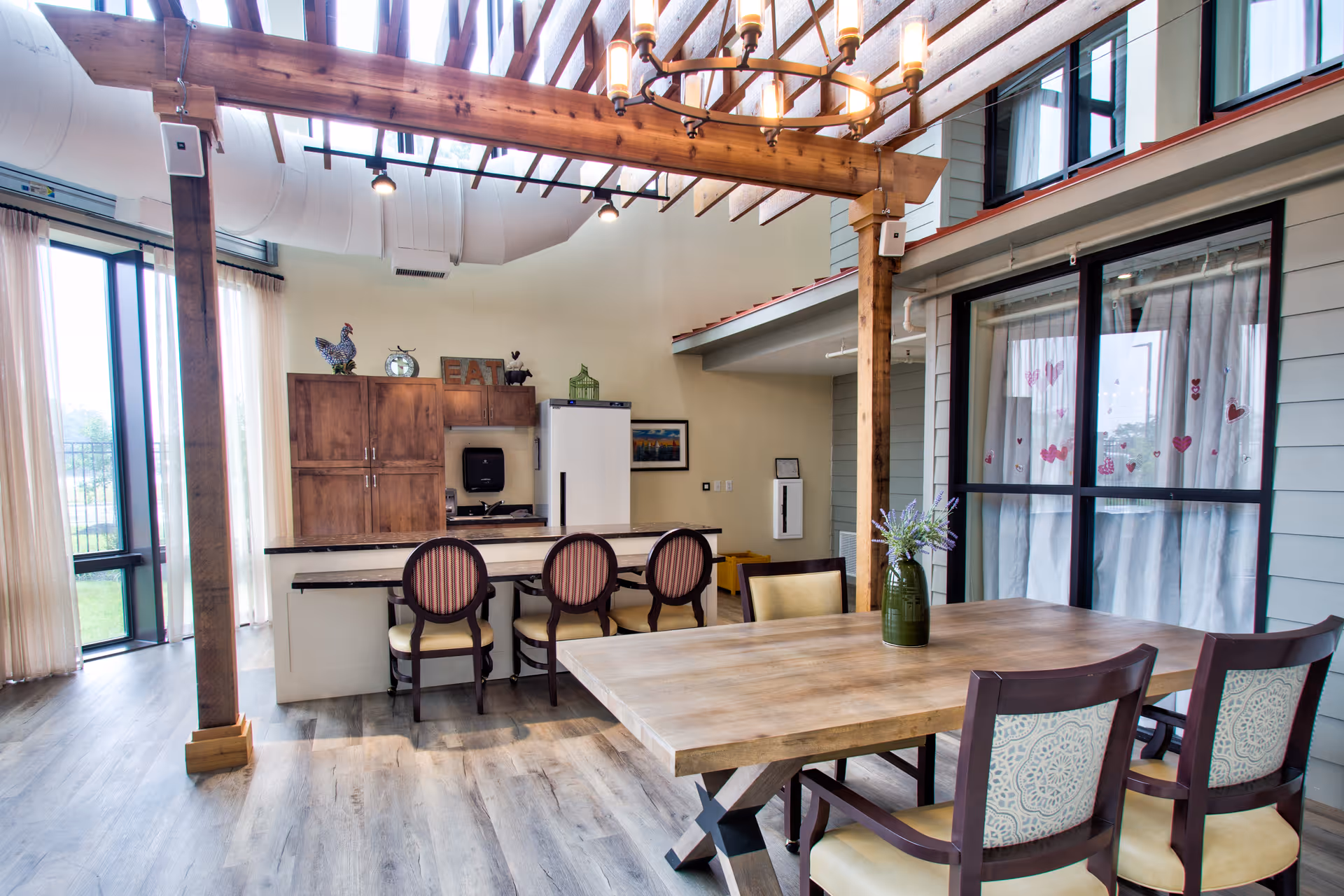 A bright and spacious dining area with a wooden table and six chairs, a kitchen island with three chairs, wooden cabinetry, a refrigerator, and large windows with sheer curtains letting in natural light. The ceiling features exposed wooden beams and a chandelier.