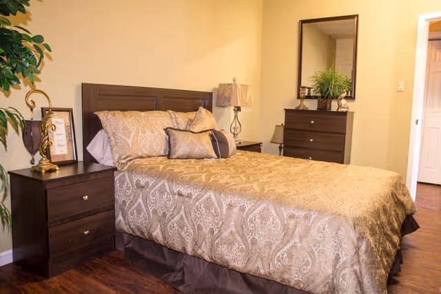 A neatly made bed with a patterned beige and brown bedspread and multiple pillows in a bedroom. The room features dark wooden furniture including a headboard, two nightstands with lamps, and a dresser with a mirror and a plant on top. The walls are painted light yellow and the floor is wooden.