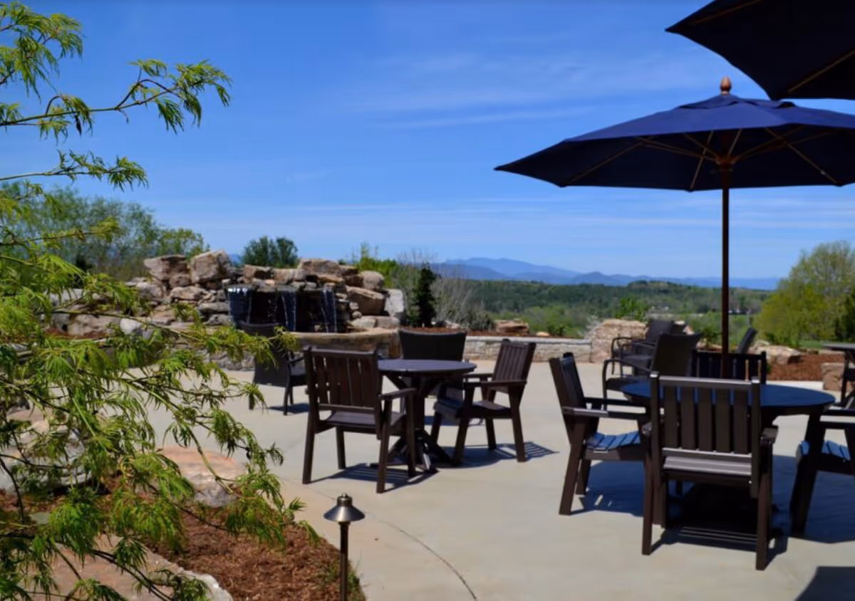 Outdoor patio area with several dark wooden tables and chairs, large blue umbrellas providing shade, a stone water feature with small waterfalls, and a scenic view of distant mountains and greenery under a clear blue sky.