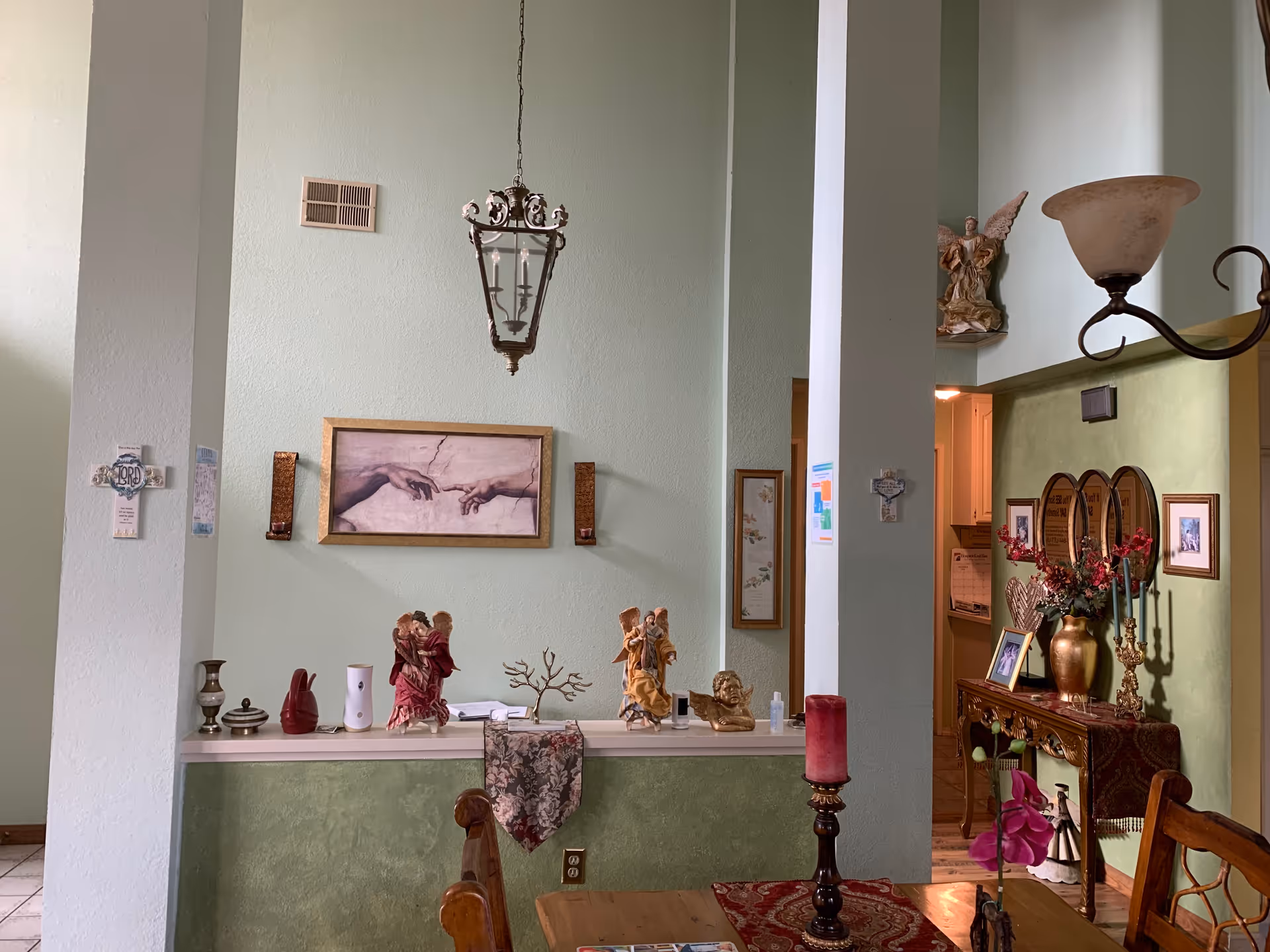 Interior view of a living room area with light green walls, decorative angel figurines on a ledge, a framed artwork of two hands reaching towards each other, a hanging lantern-style light fixture, and a wooden dining table with a red candle and floral centerpiece. There is also a side table with framed photos, flowers, and decorative items.