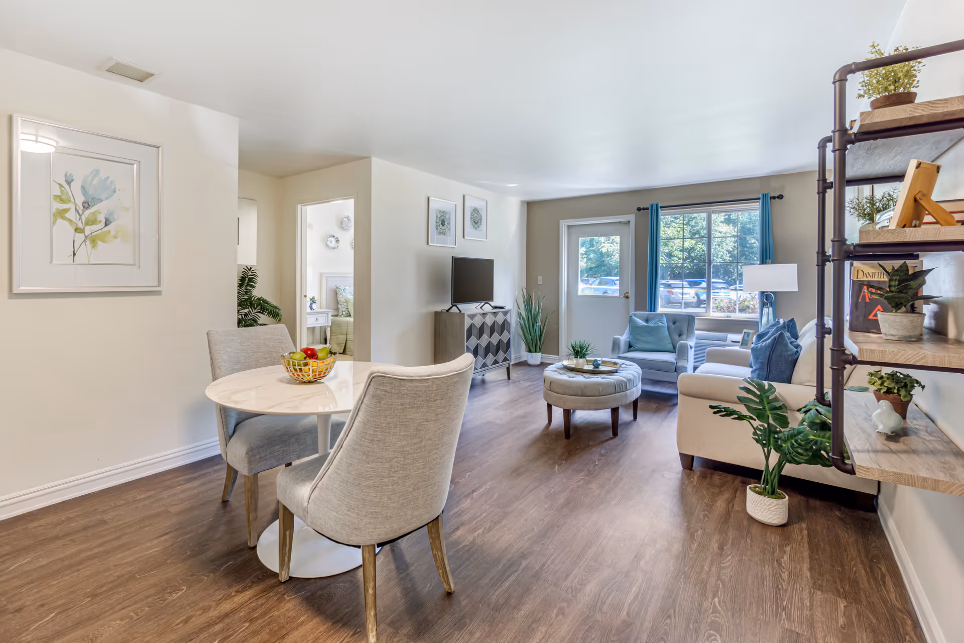 A bright and modern living area in a senior living facility featuring a small round dining table with two upholstered chairs, a cozy seating area with a sofa, armchair, and round ottoman, a TV on a patterned cabinet, and a shelving unit with decorative plants and books. A door and window with blue curtains provide natural light and a view of parked cars outside.