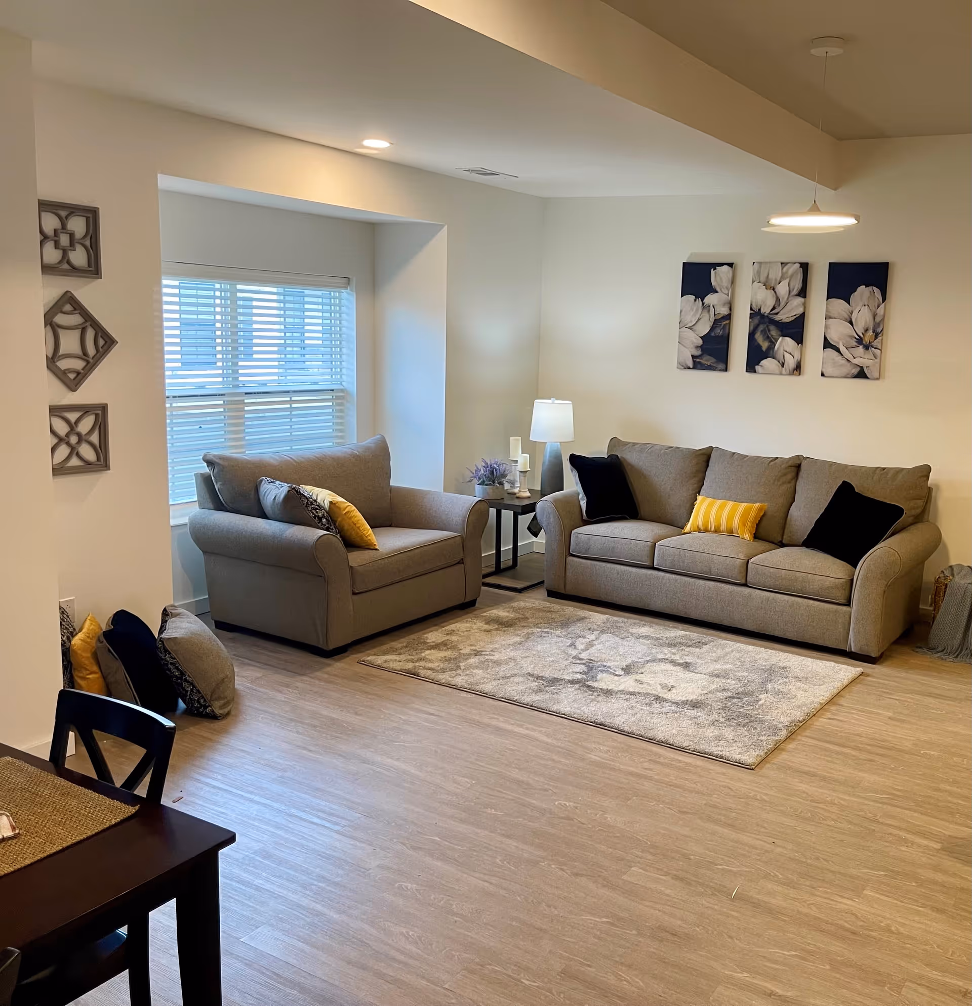 A cozy living room with two gray sofas adorned with black and yellow pillows, a beige patterned rug on a wooden floor, a side table with a lamp and decorative items, three floral paintings on the wall, and a window with white blinds letting in natural light.