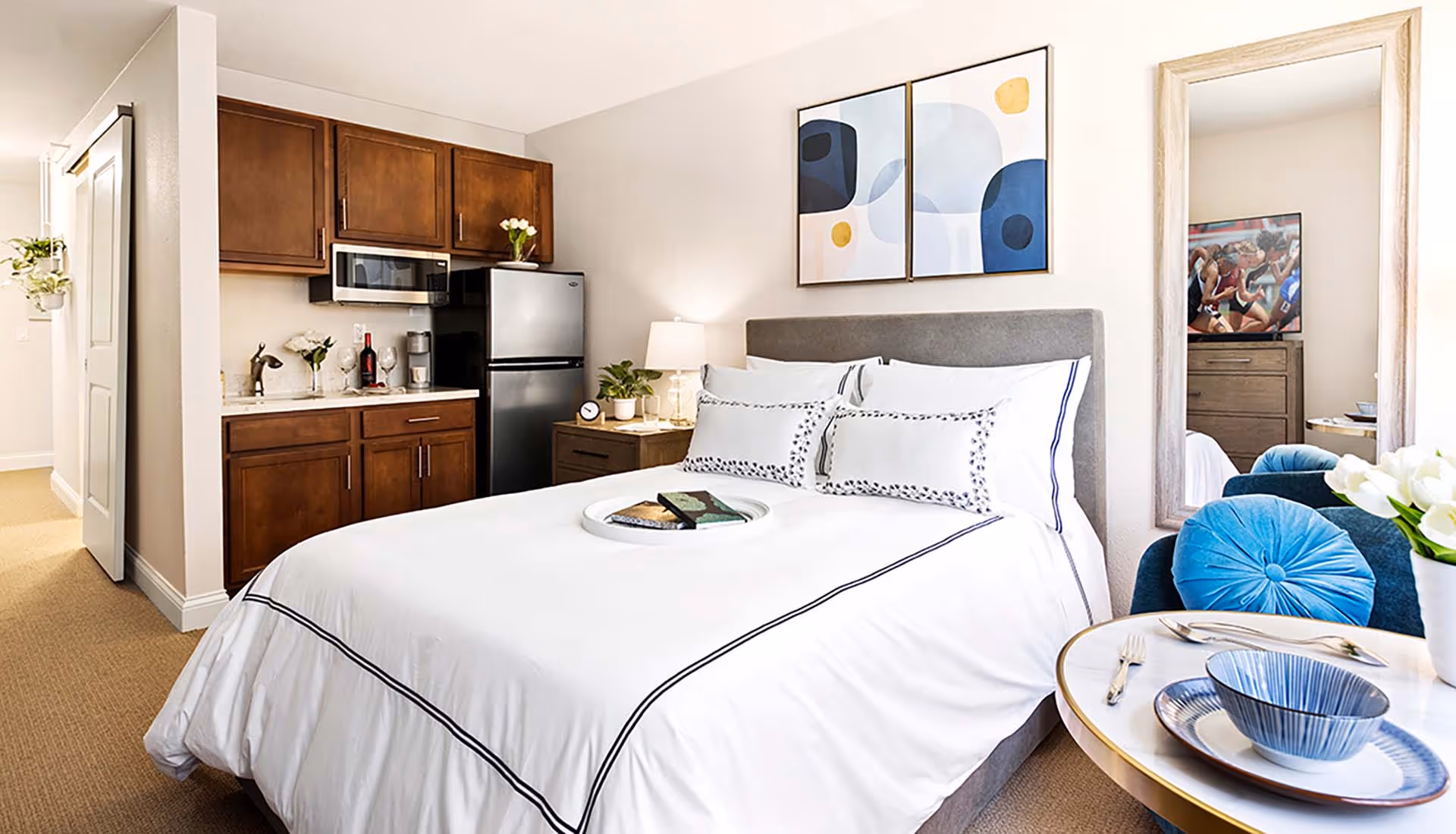 A bright and cozy senior living studio apartment featuring a neatly made bed with white linens and decorative pillows. To the left, there is a small kitchenette with wooden cabinets, a microwave, and a refrigerator. A bedside table with a lamp and a plant is next to the bed. On the right side, a round table is set with blue and white dishes and silverware, accompanied by a blue cushioned chair. A large mirror hangs on the wall, reflecting part of the room and a dresser with a TV showing a sports event. Above the bed, there are two abstract art pieces with blue, white, and yellow tones.